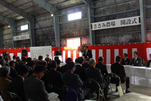 ささみ農場 開設式と書かれた幕の前でスーツ姿の男性がマイクで座っている20名ほどの人たちに話をしている写真